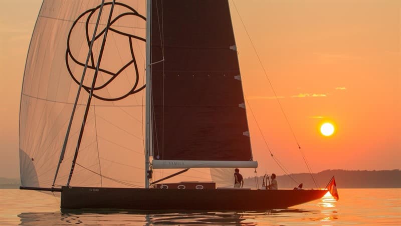 Sailboat silhouetted beside the setting sun on calm water.