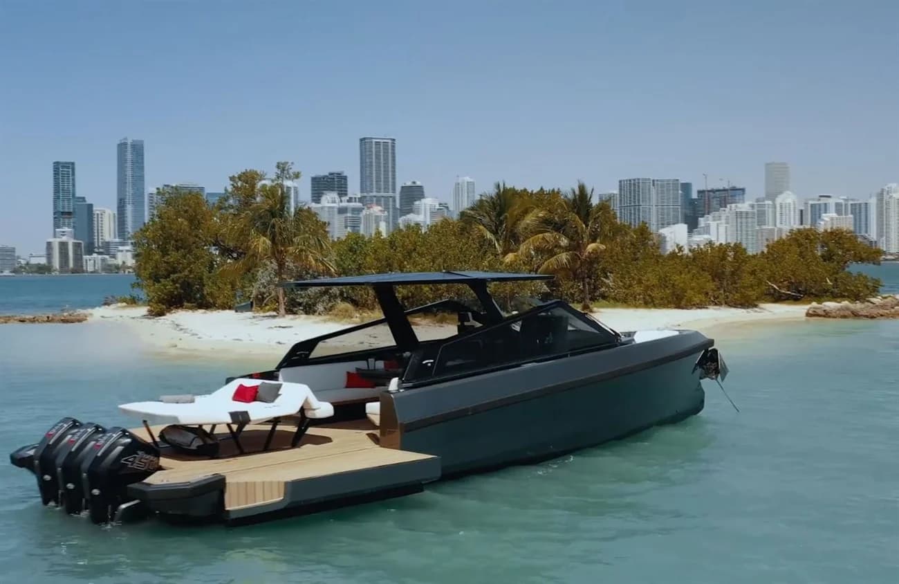 Dark motor yacht anchored near a sandy shore with a city skyline beyond.
